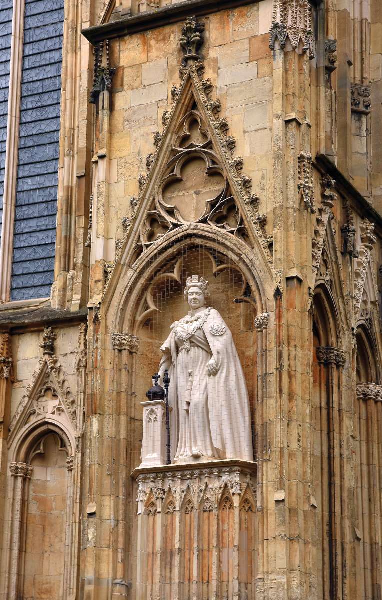 York Minster – A hatalmas homlokzat tele van csúcsíves szoborfülkékkel, ezek többségéből azonban hiányzik a szobor. Ám az egyik fülkében van egy, mely fehér színével elüt a homlokzat sárgás jellegétől. Az alak rögtön ismerősnek tűnik. Pont úgy néz ki, mint II. Erzsébet királynő – és igen, valóban ő az. Eddig csak régóta halott történelmi személyeket, mitikus figurákat, angyalokat, szenteket, stilizált alakokat láttam ilyen pozícióban. És tessék, most itt van az „éppcsak elhunyt” II. Erzsébet. A szobor eredetileg a királynő platinajubileumára (70 éves uralkodási évfordulójára) készült, és a királynő még akár láthatta is egy hónappal a halála előtt, ám felállítására már csak két hónappal a temetése után került sor, 2022 novemberében. A kéttonnás, mészkőből faragott szobor ünnepi díszben, határozott arckifejezéssel, országalmával és jogarral ábrázolja a királynőt Fotó: Telbisz Tamás